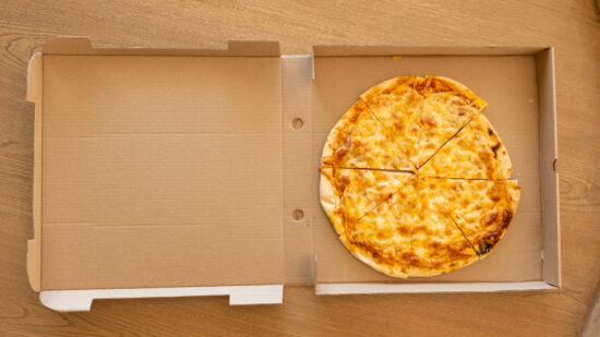 Round cheese pizza is resting in open corrugated box on wooden tabletop, showing charred crust
